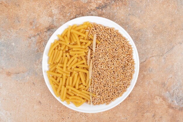 A white plate of grains and wheat with raw macaroni on marble background. High quality photo