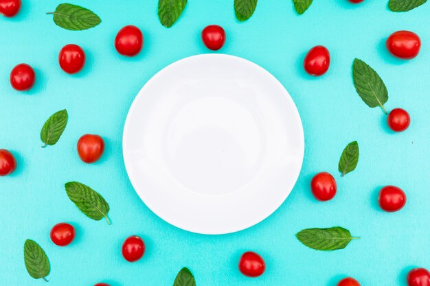 White plate surrounded with red cherry and mint top view on blue surface