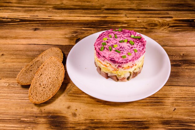 Белая тарелка с традиционным русским новогодним салатом из сельди под шубой и ржаным хлебом на деревенском деревянном столе