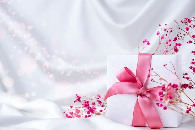 흰색 실크 직물 배경에 분홍색 리본과 작은 분홍색 꽃이 있는 흰색 선물 상자. 휴일 인사말 카드입니다. 공간을 복사합니다. 잡초 생일 발렌타인 어머니 여자의 날 프리미엄 사진