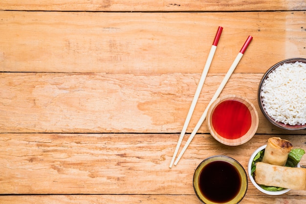 White rice bowl; spring rolls and sauces with chopsticks on wooden desk