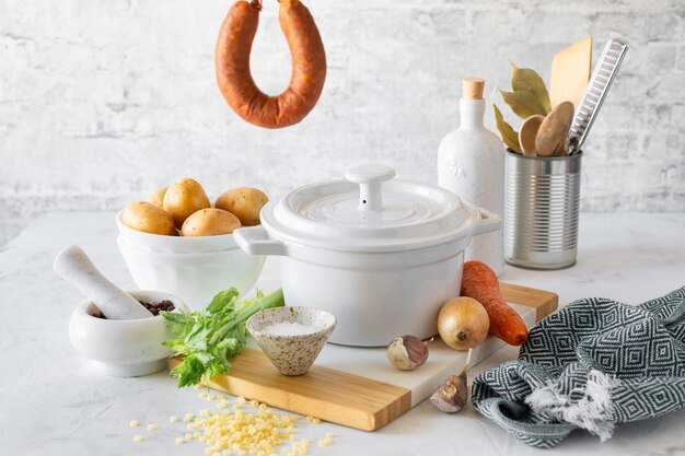 A white saucepan and ingredients for making Minestrone soup with smoked sausage on a white table Italian soup with pasta and seasonal vegetables