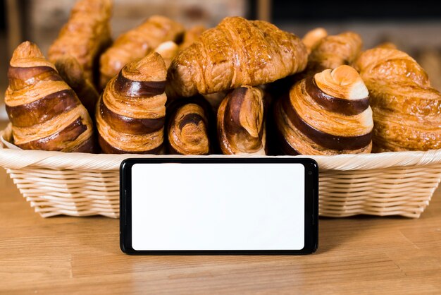Белый экран мобильного телефона рядом с корзиной, полной запеченного круассана на деревянном столе