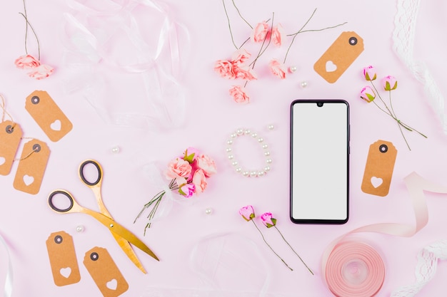 White screen display mobile phone with ribbons; roses; tags and pearl on pink backdrop