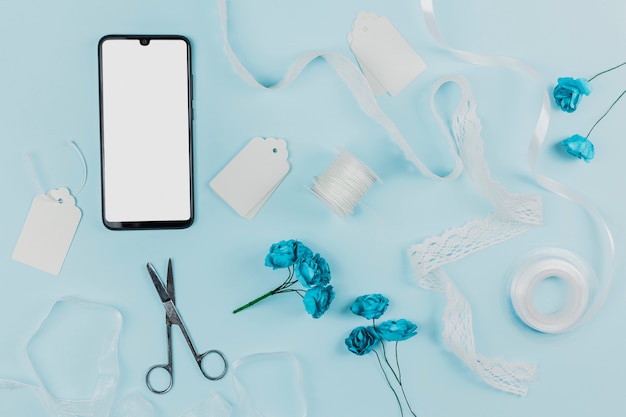 White screen smartphone with tags; spools; ribbon; scissor and artificial roses on blue background