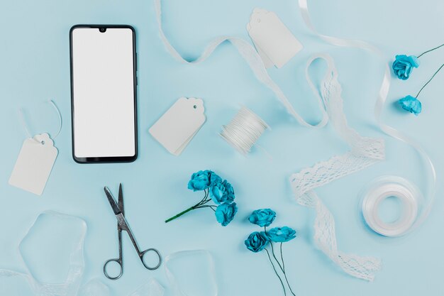 Белый экран смартфона с метками; бобины; ленты; ножницы и искусственные розы на синем фоне