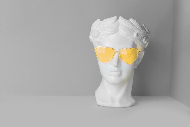 White sculpture of an antique head in yellow glasses with hearts. on a geometric background of two colors.