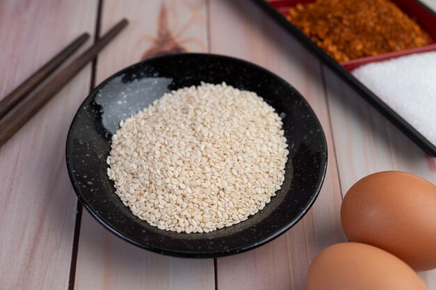 Семена белого кунжута находятся на черной тарелке, в комплекте с яйцами и палочками для еды.