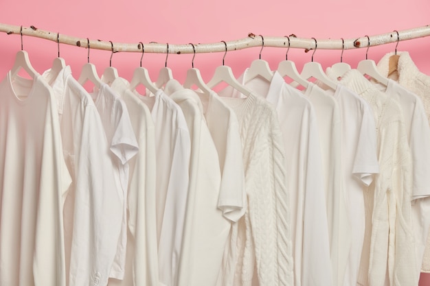 White solid clothes hanging in one row on wooden racks against pink background. Big choice for women in retail shop.