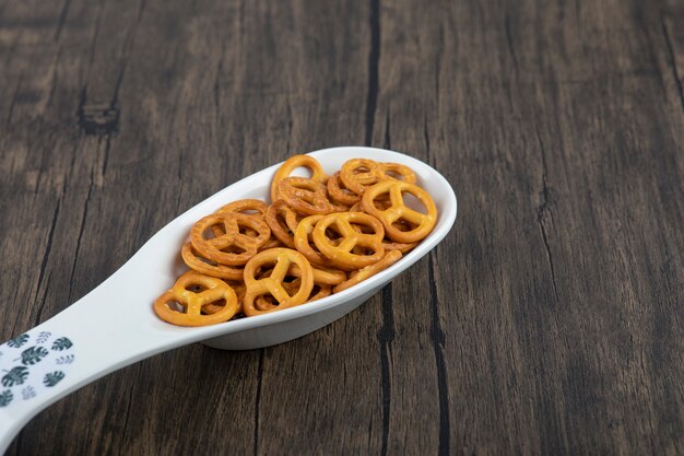 White spoon full of mini pretzel crackers placed on a wooden table .