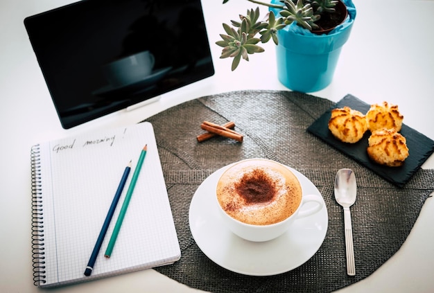 自家製カプチーノ、コーヒーとミルク、ペストリーと朝食用の白いテーブル。タブレットと背景の植物 Premium写真
