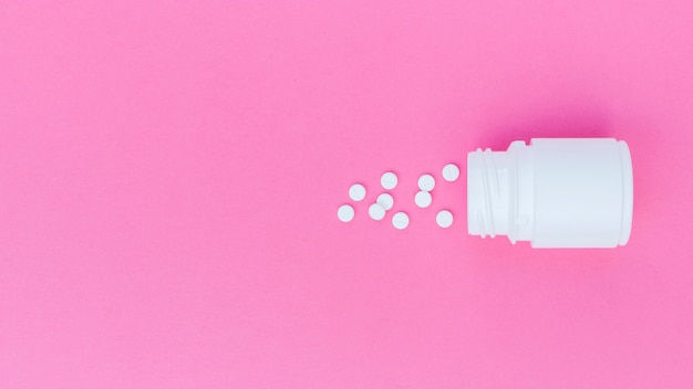 Белые таблетки пролились из пластиковой бутылки на розовом фоне