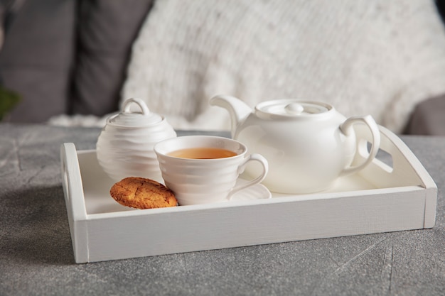 White tea set and cakes on wooden grey table. White wooden tray with cups, teapot and garland lights.