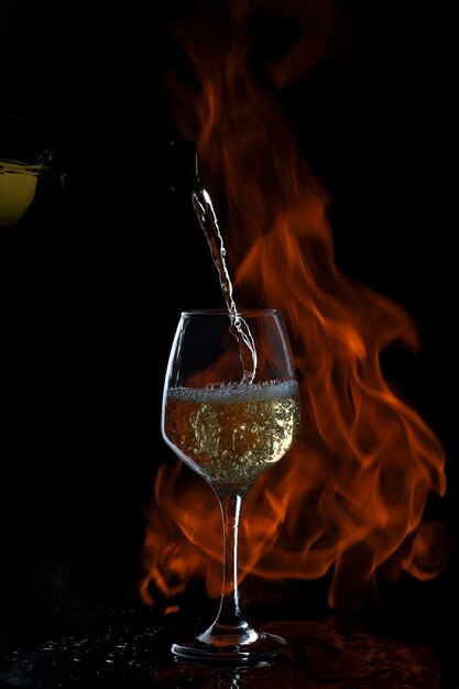 white wine is being poured to glass with long stem in dark backgrond with fire