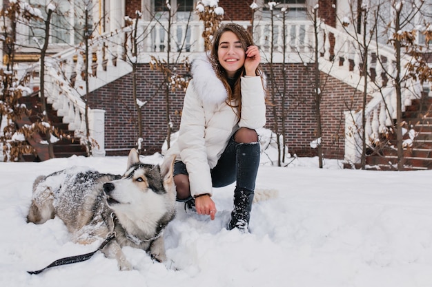 Donna bianca con un sorriso incredibile in posa con il suo cane durante la passeggiata invernale in cortile. la foto all'aperto della signora allegra indossa pantaloni di jeans strappati seduti sulla neve con un husky pigro.