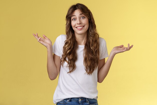 Who cares have fun. Carefree indifferent outgoing happy young woman shrugs raise hands sideways clueless unbothered apathic to topic uninterested smiling broadly careless yellow background