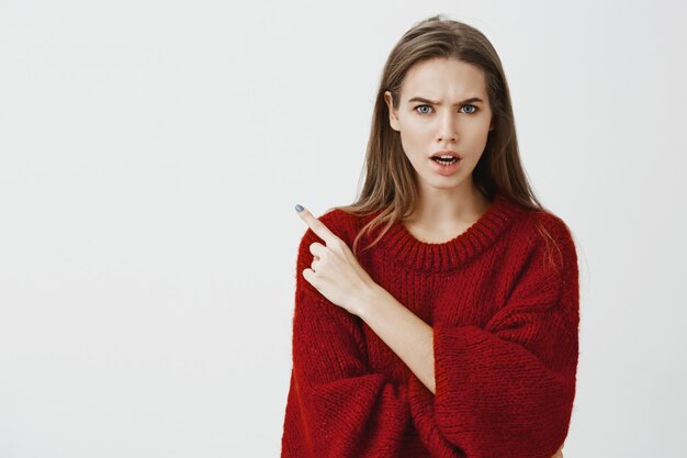 Who invited that drunk guy to party. Studio shot of displeased angry young woman in red loose sweater, frowning and arguing with friend, pointing at upper left corner 