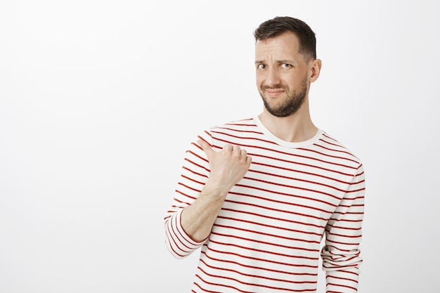 Free photo who invited that guy. portrait of displeased doubtful attractive male in striped pullover, grimacing from disgust and pointing left with thumb, being unsure