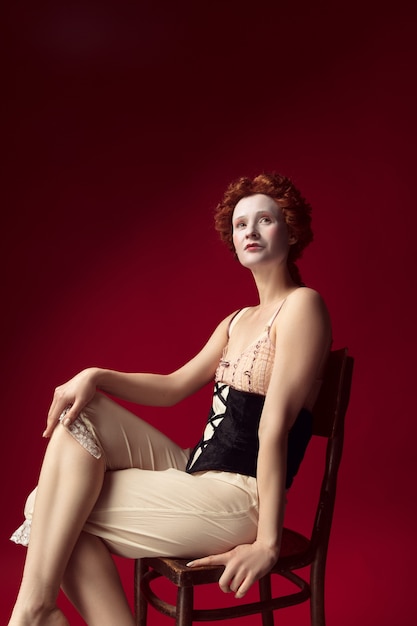 Free photo who is in control. medieval redhead young woman as a duchess in black corset and night clothes sitting on the chair on red wall. concept of comparison of eras, modernity and renaissance.