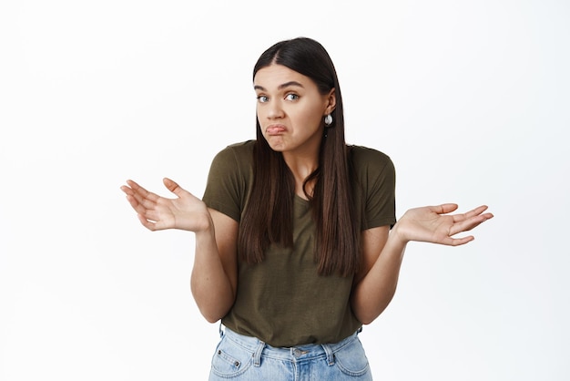 Free photo who knows cant tell clueless and confused young brunette woman shrugging spread hands sideways and look uninvolved know nothing standing over white background