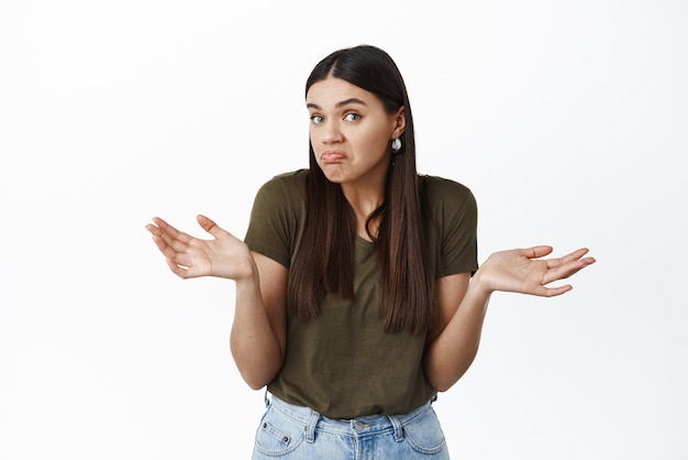 Foto gratuita chissà non può dirlo la giovane donna bruna senza tracce e confusa che scrolla le spalle allarga le mani di lato e sembra non coinvolta non sa nulla in piedi su sfondo bianco