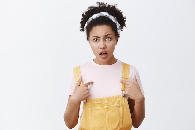 Who, me. Portrait of puzzled and confused displeased cute female with dark skin in headband over hair and yellow trendy overalls, being insulted and disappointed, pointing at herself over grey wall