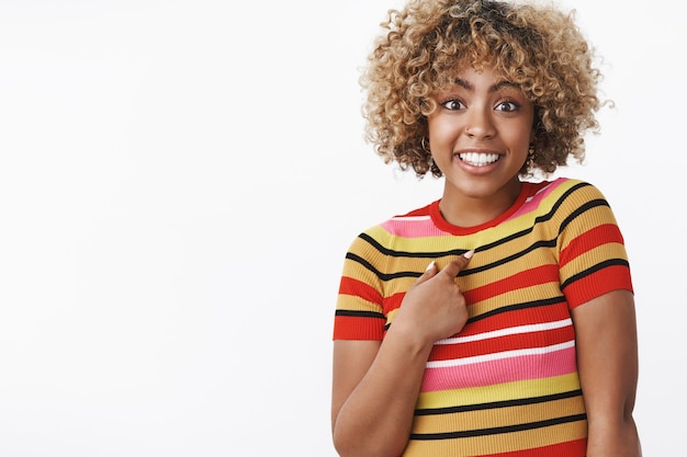 Who me. Portrait of surprised cute and carefree charming african-american girl pointing questioned and excited at herself as being mentioned or picked smiling broadly posing over white wall