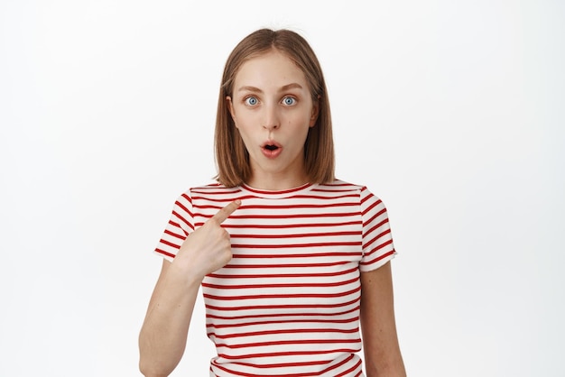 Who me Surprised blond girl student looking in disbelief and awe at camera pointing at herself being chosen winning something achieve prize staring shocked white background