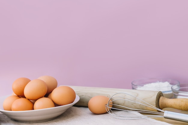 Цельные коричневые яйца на тарелке; скалка и венчики на розовом фоне