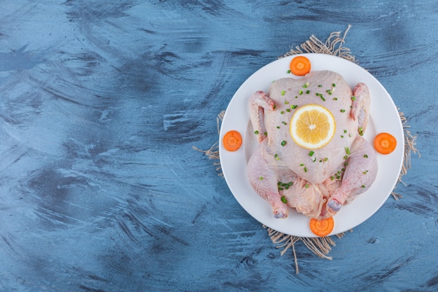 Whole chicken, lemon and sliced carrots on a plate on a burlap, on the blue background.