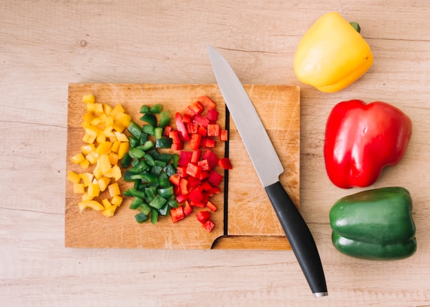 Whole and chopped red; green; yellow bell peppers on chopping board with sharp knife against wooden backdrop