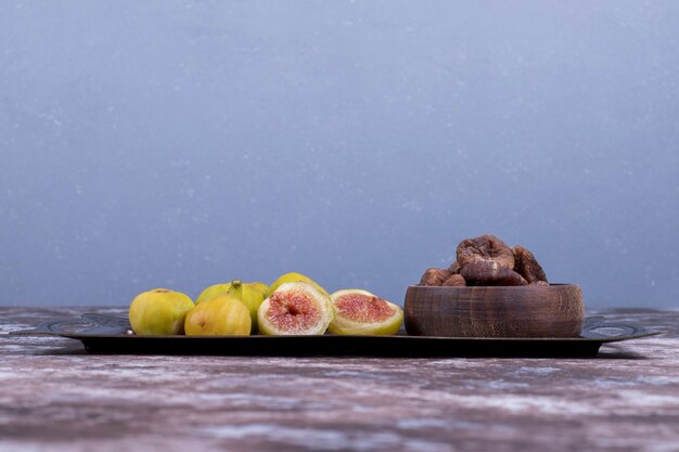 Whole, dry and sliced figs in a metallic tray and in a wooden cup on blue.