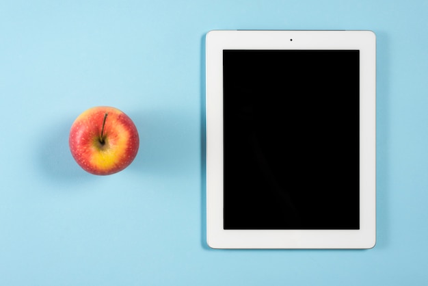 Целое красное яблоко возле цифрового планшета с пустым экраном на синем фоне