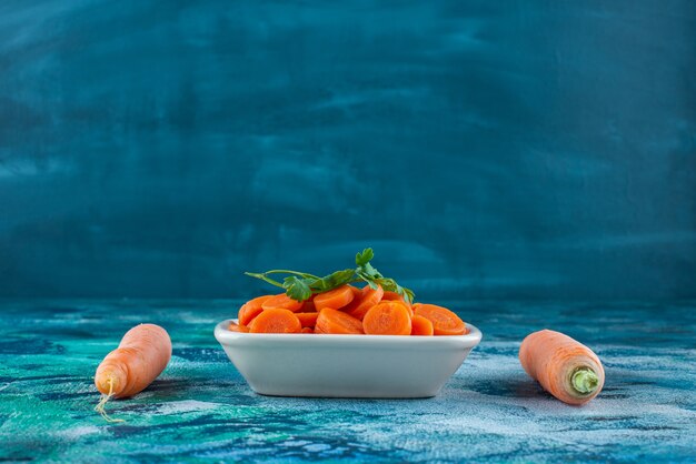 Целую морковь с петрушкой нарезать ломтиками в миске, на синем столе.