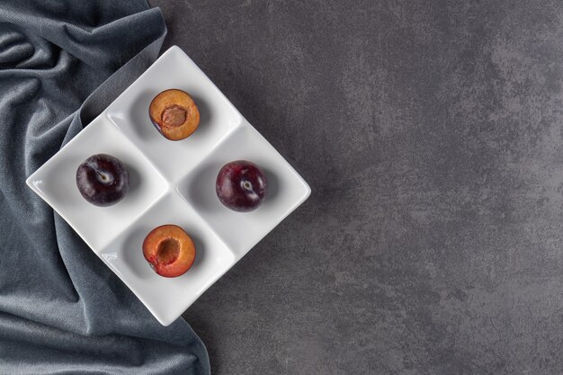 Whole and sliced juicy red plum fruits placed on a white plate.