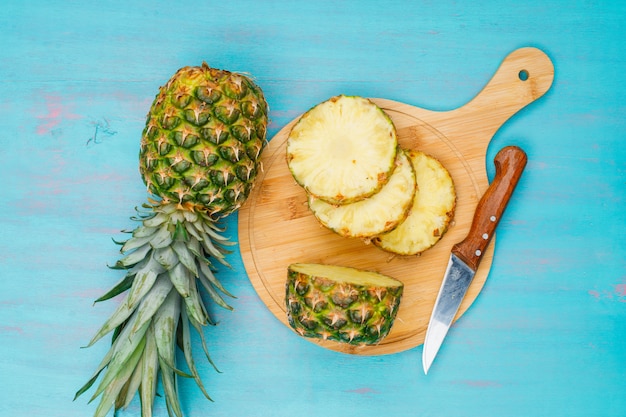 Free photo whole and sliced pineapple with a fruit knife in a cutting board on blue cyan, flat lay.