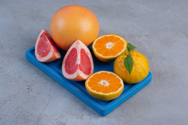 Целые и нарезанные мандарины и грейпфруты на маленьком синем подносе на мраморном фоне.