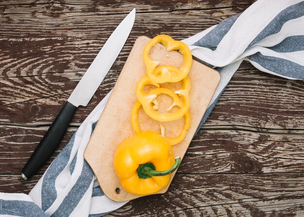 Whole and slices of yellow bell pepper on chopping board over the stripes cloth with sharp knife against wooden desk