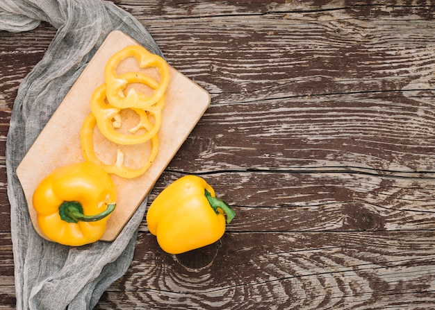 Whole and slices of yellow bell peppers on chopping board over the grey textile against wooden desk