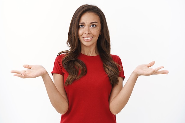 Why so serious. Questioned surprised young cute brunette female with freckles in red t-shirt cannot understand what happening, shrugging raise hands up clueless, smiling concerned