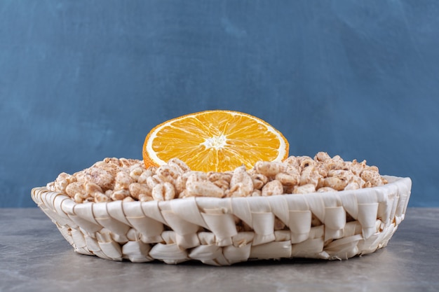 Un cesto di vimini di cereali per la colazione di riso croccante sano con una fetta di frutta arancione.