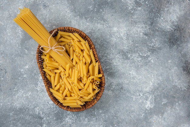 Wicker basket of raw spaghetti and penne pasta on marble surface.