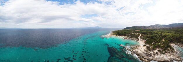 エーゲ海の岩の多い海岸の広角ショット、周囲の緑、茂みと木々、丘と山、波のある青い水、ギリシャのドローンからの眺め