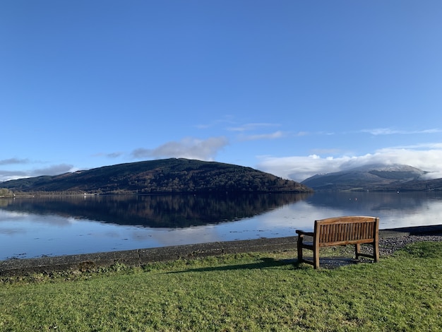 Wide angle shot of a wooden bench on a green field in front of water and a mountain