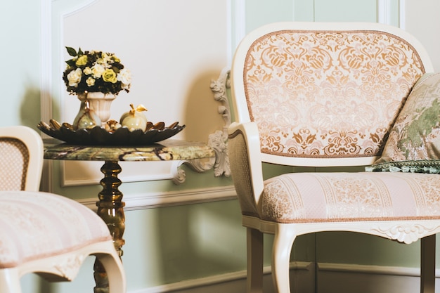 Wide closeup shot of a flower bouquet in a vase on a table near an elegant armchair