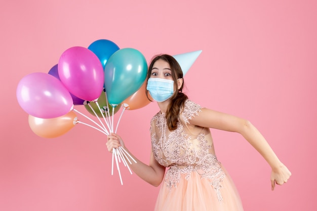 Free photo wide-eyed girl with party cap running while holding colorful balloons on pink