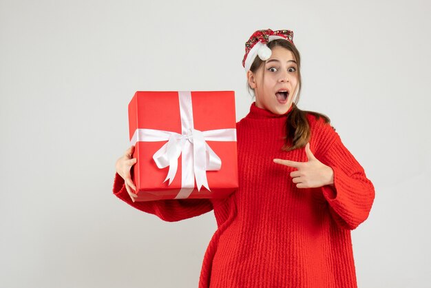 wide-eyed girl with santa hat holding present finger pointing the box standing on white