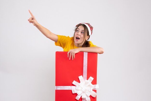 wide-eyed girl with santa hat pointing at something standing behind big xmas gift on white