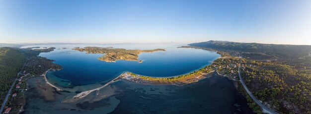 Wide shot of the Aegean sea coast with a town on the shore and island, blue transparent water, greenery around, pamorama view from the drone, Greece