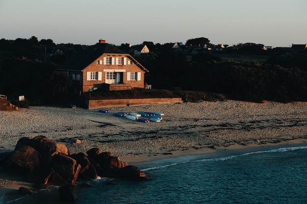 Wide shot of a brown house on a sandy seashore by the sea surrounded with rocks and trees